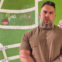 Man with a beard and tattoos wearing a brown t-shirt in front of a green wall with a map design.