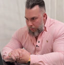 Man wearing a pink shirt with a logo, sitting and looking at something in his hands.